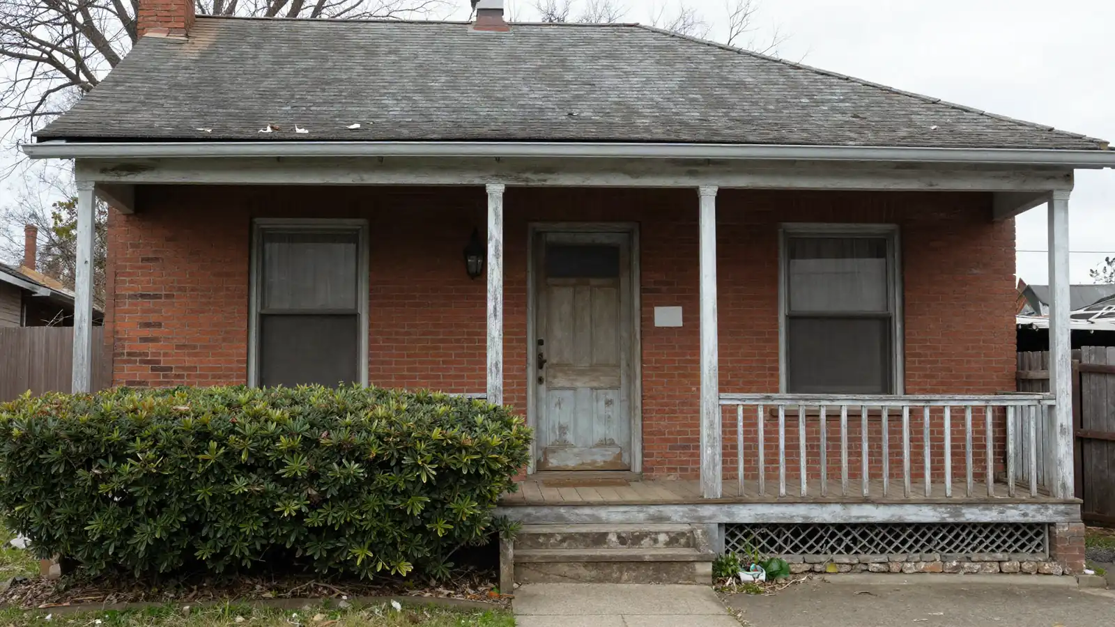 A worn older two-story brick house with an overgrown hedge and a weathered porch on an overcast day