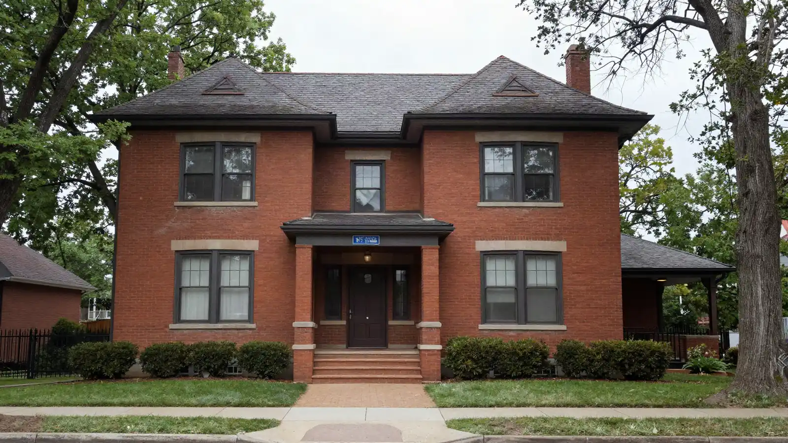 A large early 1900s brick house with a tile roof and wide front steps behind old trees