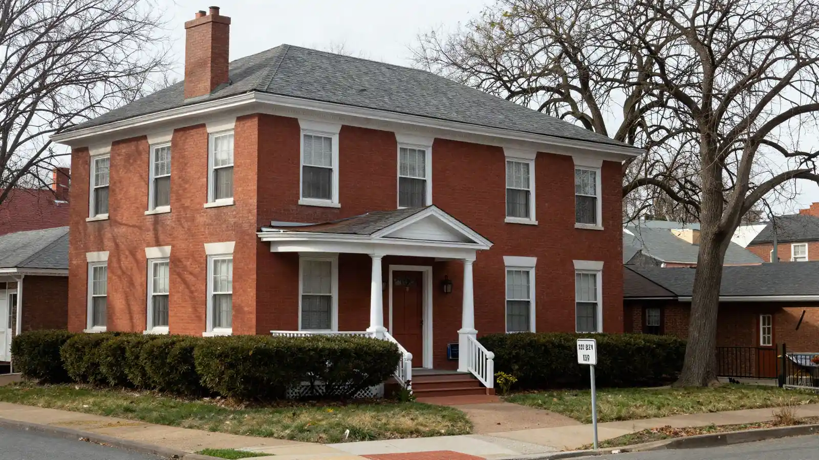 A 1920s red brick house with white trim, a small front porch and trimmed hedges in morning light