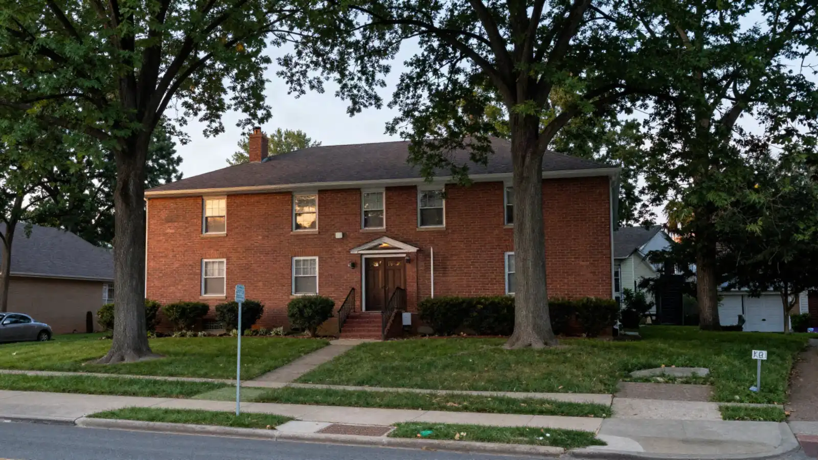 A brick split-level house on a sloped lot with steep front steps and big trees in early evening light