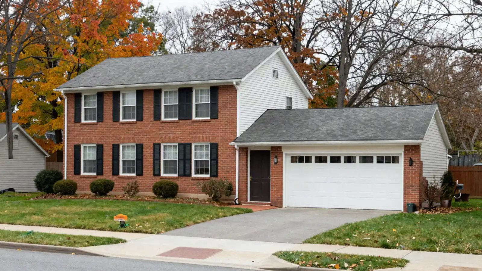 A 1970s two-story brick and siding colonial with black shutters and an attached garage in autumn