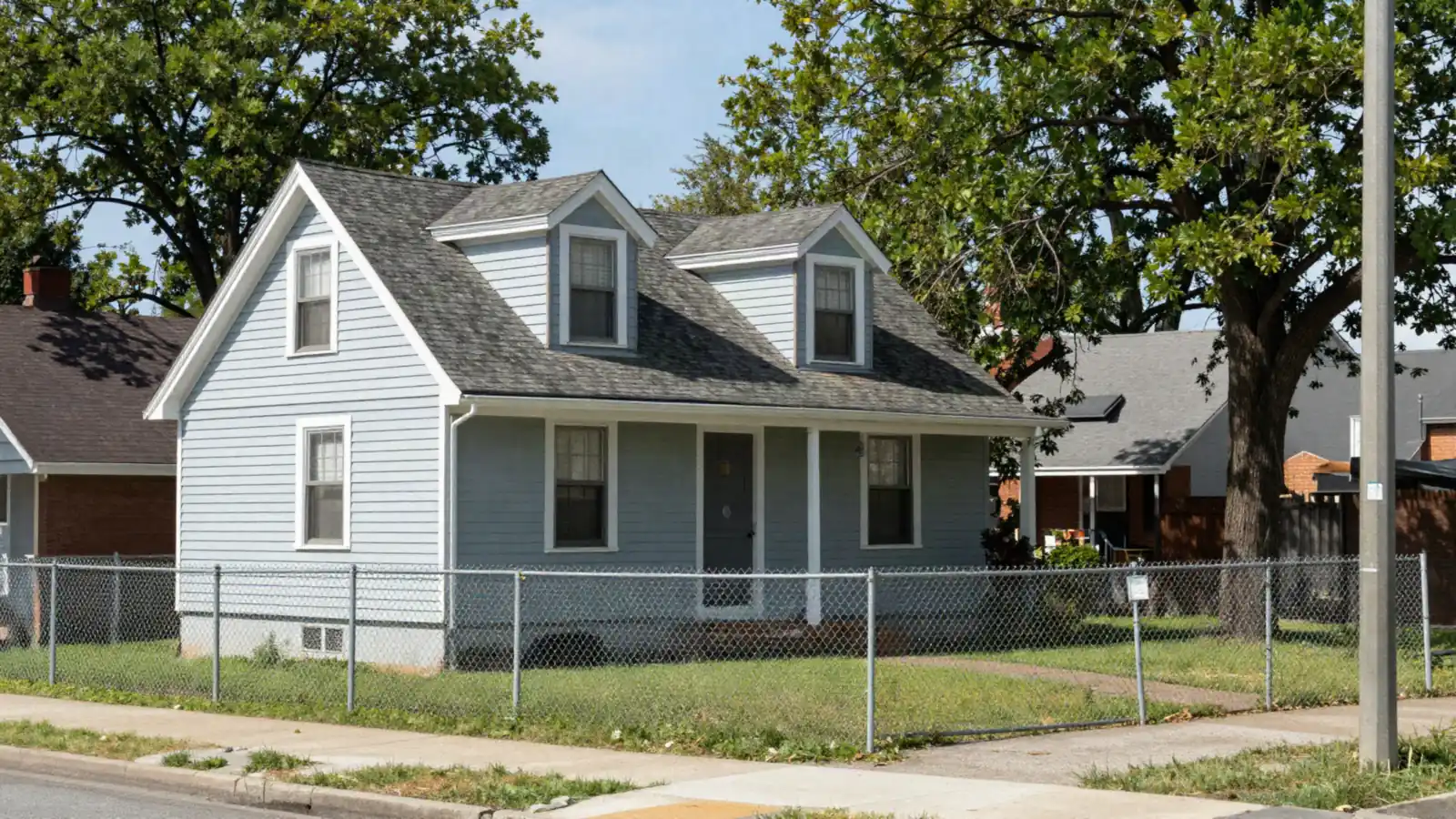 A small 1940s frame Cape Cod house with dormers and a chain-link fence in bright daylight