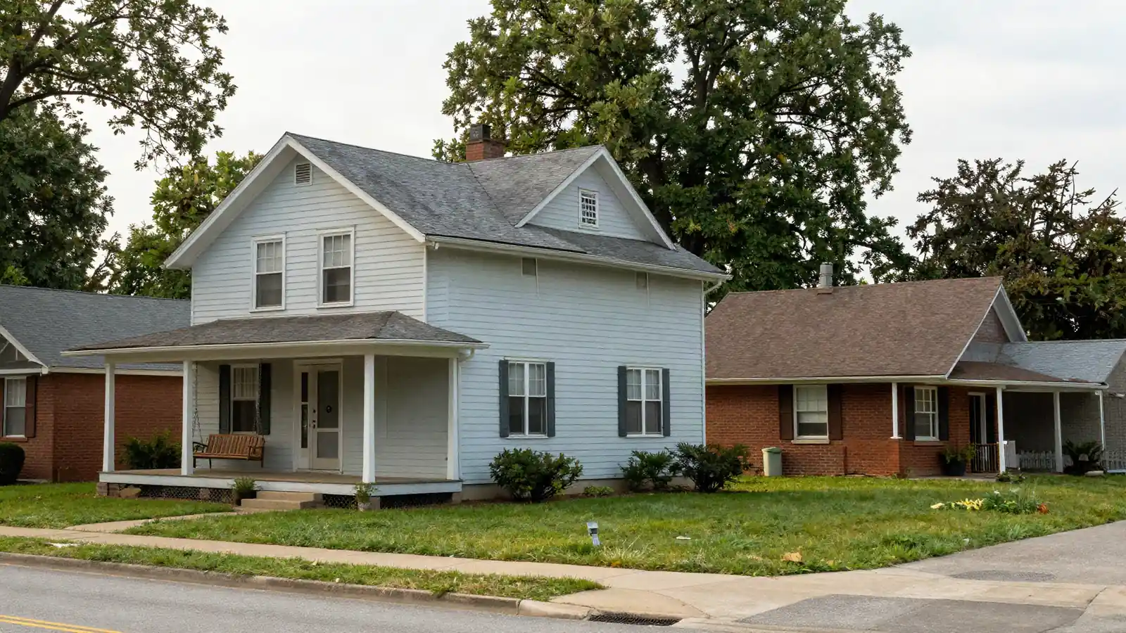 An older frame house with a porch swing beside a 1950s brick ranch on a tree-lined street