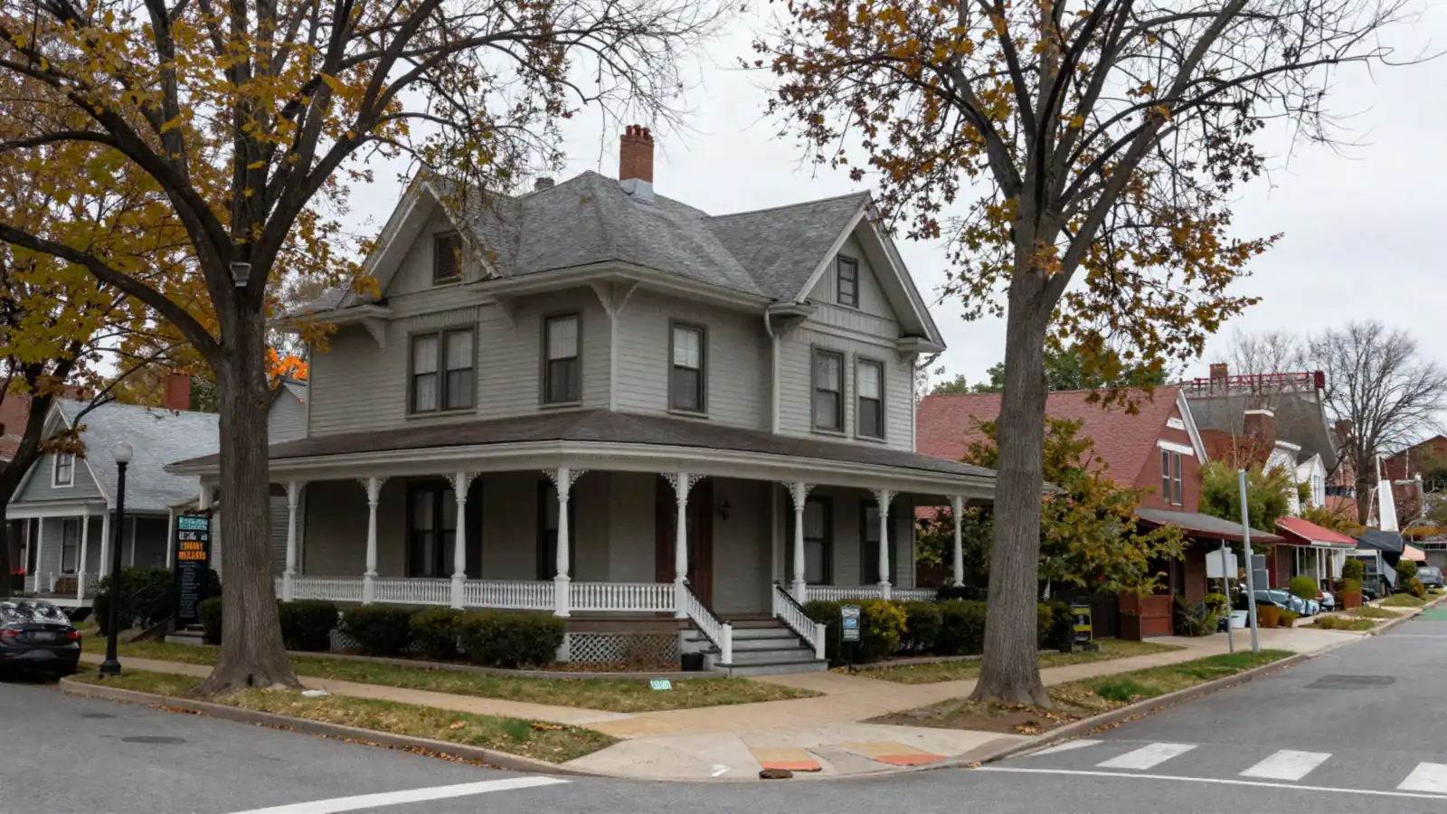 A large older house with a wraparound porch and mature trees on a wide street in autumn