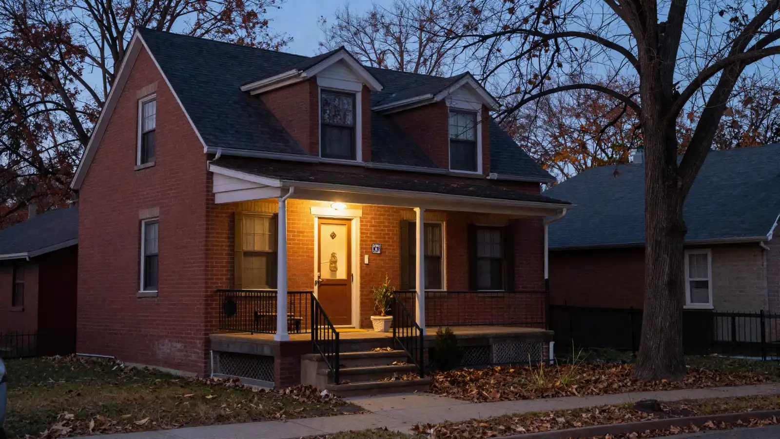 A modest two-story brick house on a hillside street at dusk with the porch light on and autumn leaves on the steps
